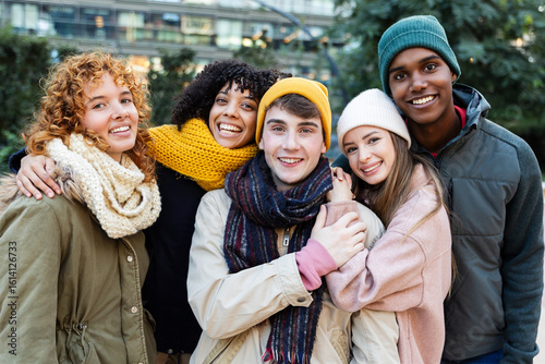 Young happy group of diverse friends in warm clothes smiling at camera standing together outside. Portrait of millennial people enjoying winter vacation. Friendship and youth concept.