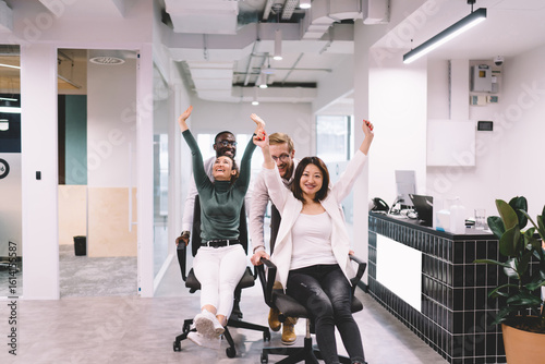 Multiethnic colleagues burst into laughter while racing chairs through office, capturing fun, unity, and emotional recharge in relaxed, engaging workplace environment