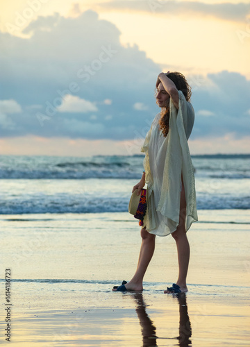 Full-body portrait of a Colombian woman standing on wet sand at the beach during sunset, holding a colorful traditional hat and looking at the horizon.