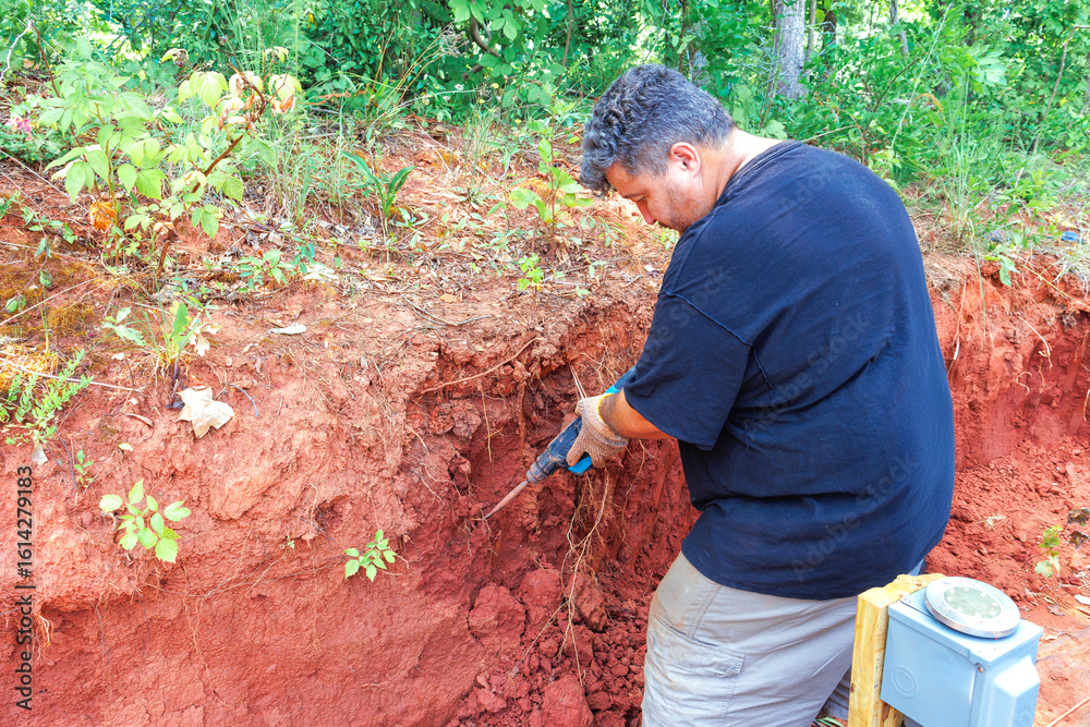 Naklejka premium Worker man uses jackhammer tools to dig remove roots from reddish soil while surrounded by greenery trees.