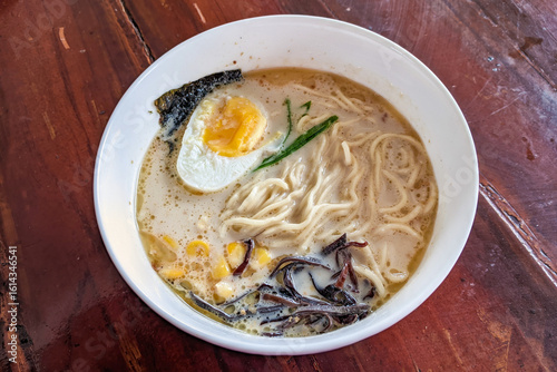 A steaming bowl of creamy ramen with noodles, a perfectly cooked egg, corn, and seaweed on a rustic wooden table.