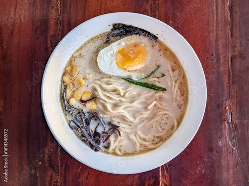 A steaming bowl of creamy ramen with noodles, a perfectly cooked egg, corn, and seaweed on a rustic wooden table.