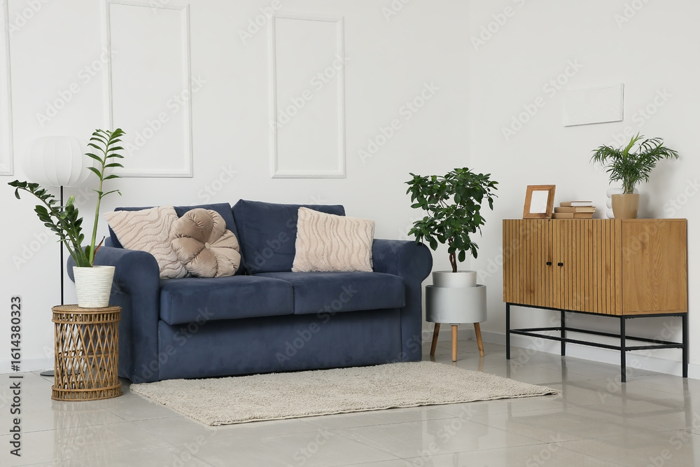 Obraz premium Cozy blue sofa with dresser, coffee table and houseplants near white wall in living room