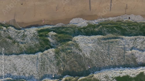 Slow motion top-down drone footage shows Atlantic Ocean waves rolling and foaming as they crash on Blouberg Beach near Cape Town, with surfers scattered near the sandy shoreline and entering water.