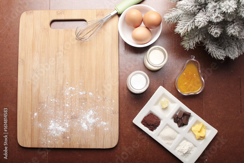 A top-down view shows baking ingredients including eggs, milk, flour, cocoa powder, chocolate, and butter laid out on a dark wooden surface next to a cutting board and a frosted pine branch