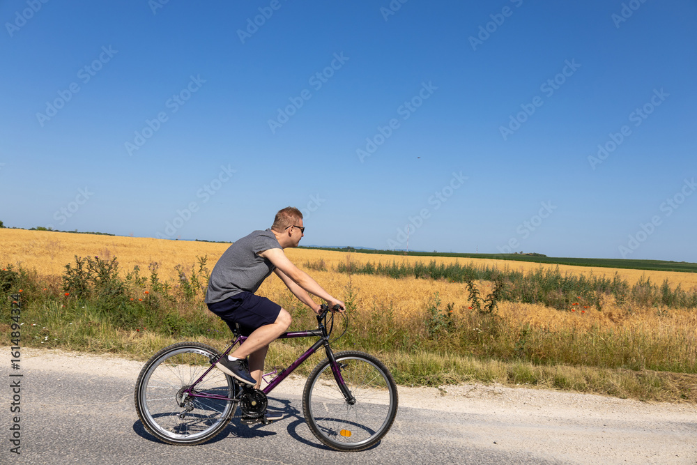 Fototapeta premium man cycling on a rural road beside a golden wheat field under summer sky