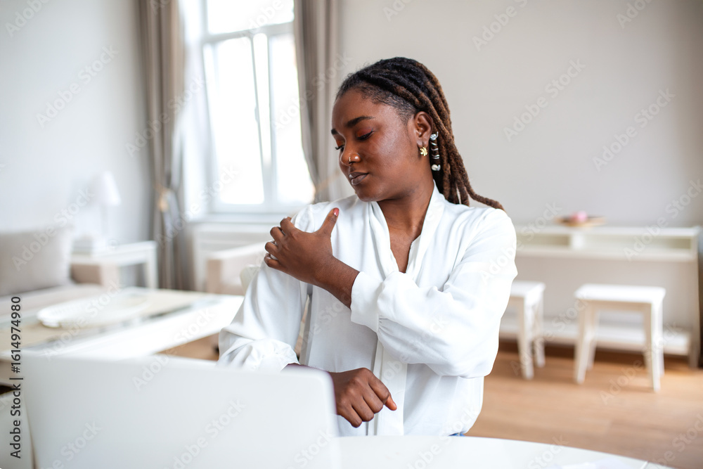 Portrait of young stressed African woman sitting at home office desk in front of laptop, touching aching shoulder with pained expression, suffering from shoulder ache after working on pc Portrait of young stressed African woman sitting at home office desk in front of laptop, touching aching shoulder with pained expression, suffering from shoulder ache after working on pc