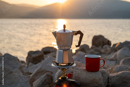 Red metal cup and moka pot on portable stove against peaceful lake and hills background at dusk, perfect scene for rustic travel, hiking break, nature lovers, coffee ritual