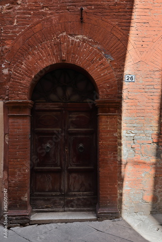 Round-arch door cut in the red brick facade of a townhouse on Via Mazzini Street where the sun casts sun and shade games. Montalcino-Tuscany-Italy-134
