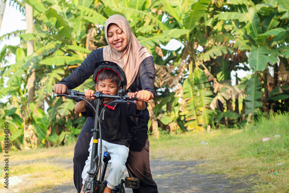 Fototapeta premium Small boy riding his bike for the first time. Young mom helping son riding a bicycle. Mom teaches her little son to ride a bicycle in the city park. Happy family moments. Time together mother and son.