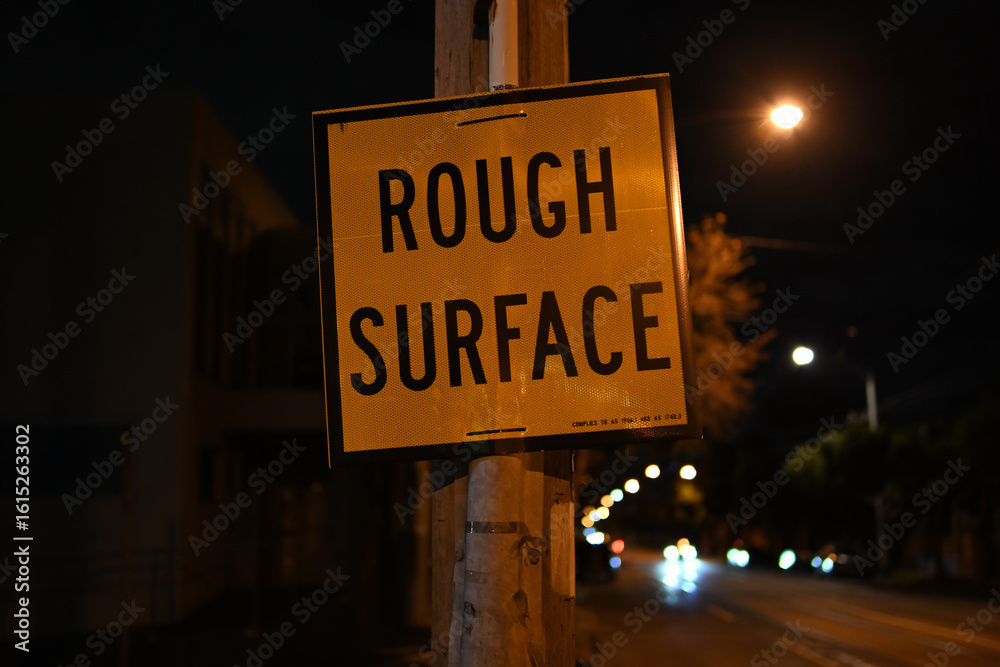 Fototapeta premium Yellow and black rough surface traffic sign, attached to a power pole, beside a road at night