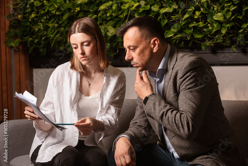 Two business colleagues discuss documents intently on a modern sofa. Professional teamwork, thoughtful expressions, stylish casual office interior with green wall decor. Businessman and assistante .