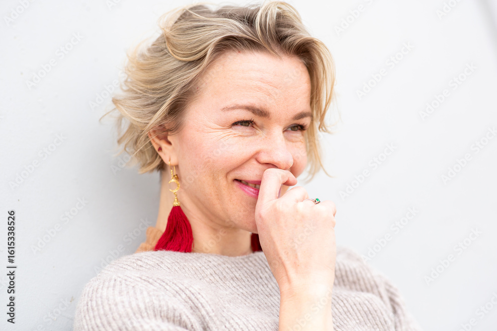 cheerful woman with short blond hair smiles playfully while leaning against a white wall cheerful woman with short blond hair smiles playfully while leaning against a white wall