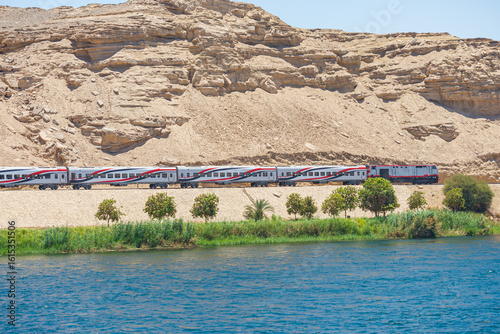 Train of the railway is running on the eastern bank of the river Nile, Egypt. Blue waters in the foreground, with green trees. Dry hill on the background.