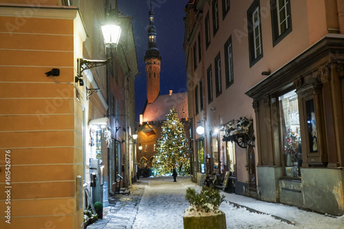 Fototapeta Naklejka Na Ścianę i Meble -  Cute small street in Tallinn old town leading up to a decorated Christmas tree and a Church in the background on a snowy festive winter evening, Estonia