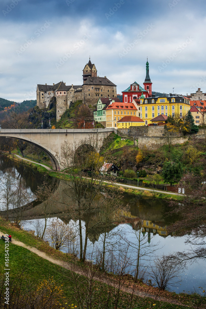 Obraz premium View of Loket Castle and colorful historic buildings in Loket, Czech Republic, with a stone bridge and river reflection on a cloudy autumn day
