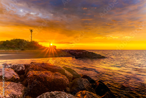 Fototapeta Naklejka Na Ścianę i Meble -  Sunrise over the lighthouse in Bibione, Italy, with golden skies, rocky shore, and calm sea – a serene coastal landscape perfect for travel and nature themes