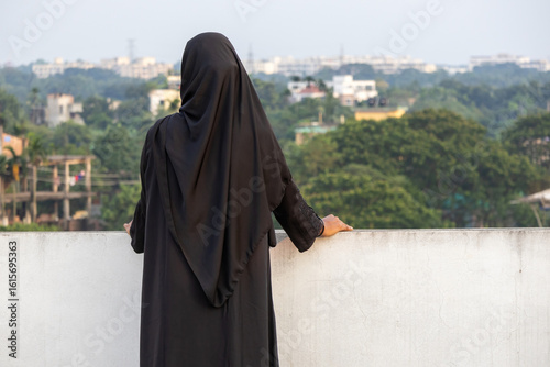 фотографія Solo Bengali Muslim woman in black hijab stands on a rooftop, back to viewer, contemplating the vast outdoor nature