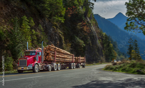 Red logging truck hauling timber through scenic forested mountains near Hurricane Ridge, Washington. Iconic American transport along curving rural highway in daylight.