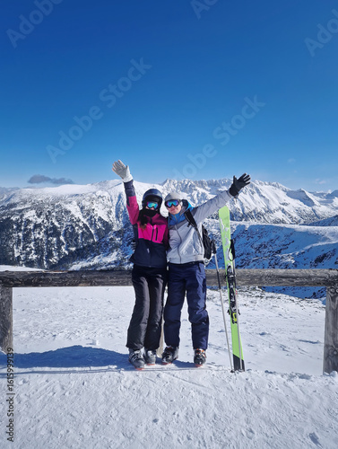 Happy couple standing on the snowy mountain top with arms raised posing as waving to camera. Skiers man and woman practice winter sports together having fun at Bansko ski resort in Bulgaria