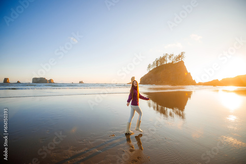 Young brunette woman running barefoot along the shoreline at sunset, Second Beach, Olympic National Park, Washington State. Scenic golden-hour beach scene with ocean and sea stacks.