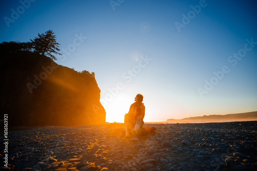 Brunette woman wearing a yellow rain jacket on Ruby Beach sitting at sunset. Scenic coastal view with sea stacks, tidepools, and warm twilight colors. Olympic National Park, Washington State.