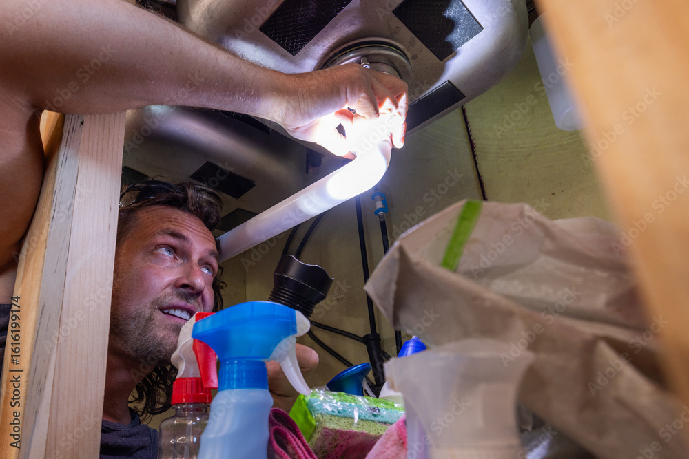 Ταπετσαρία A man uses a flashlight to inspect or repair plumbing beneath a kitchen sink, surrounded by household cleaning supplies
