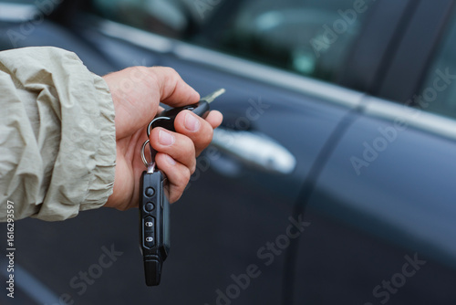 Close-up of a hand holding car keys and a key fob with remote control buttons against the background of a car. Concept of car ownership and rental.