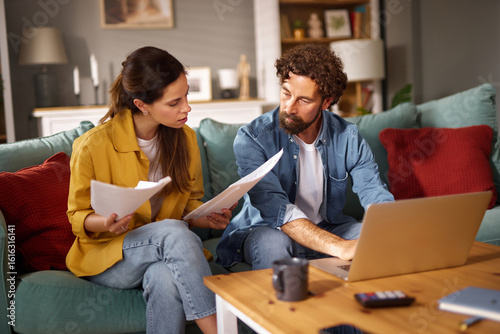 Принти на полотні Two individuals are seated on a couch, focused on financial documents and a laptop, engaged in a serious conversation about their financial issues