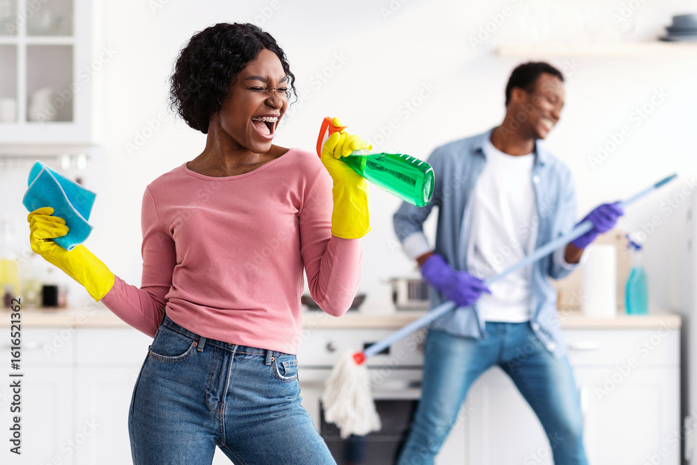 Ταπετσαρία Joyful black lady with dust cloth using spray as microphone, singing while cleaning kitchen with her cheerful boyfriend with mop