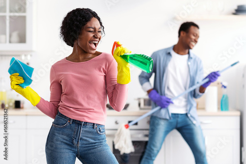 Bilde på lerret Joyful black lady with dust cloth using spray as microphone, singing while cleaning kitchen with her cheerful boyfriend with mop
