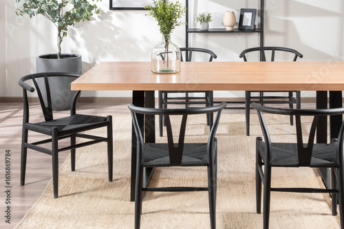 Spacious Dining Area with Sturdy Beechwood Table  Black Weave Chairs on Textured Beige Rug  Large Green Plant in Matte Charcoal Pot  Clear Vase with Green Foliage  Illuminated by Ambient Natural Light