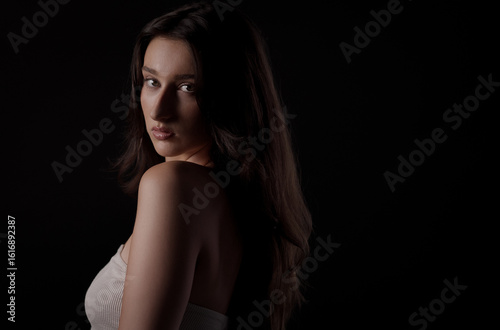 Studio portrait of beautiful young woman with long brown hair looking over shoulder on black background