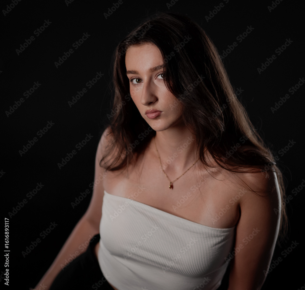 Young woman with long brown hair wearing white top and necklace posing in studio with black background Young woman with long brown hair wearing white top and necklace posing in studio with black background