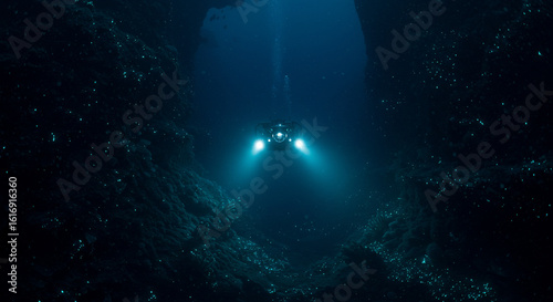 A submersible with bright lights descends into the deep blue ocean with dark rocky surroundings