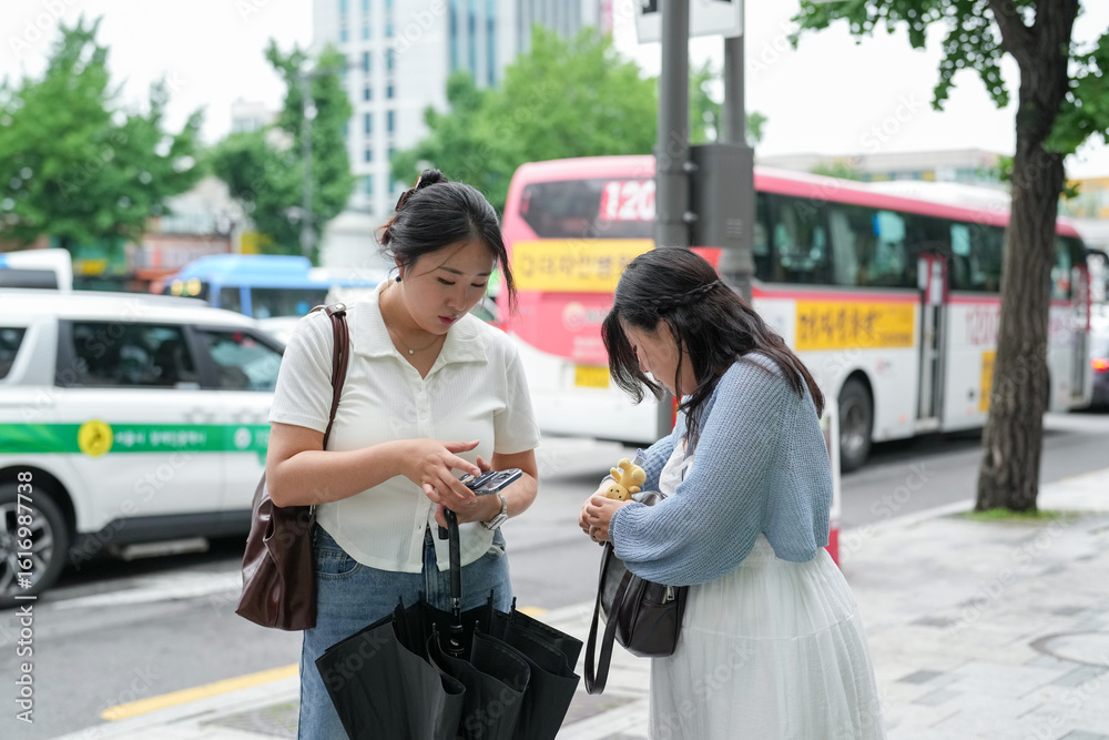 Φωτογραφία On a busy boulevard of honking cars and bustling crowds, two Korean women in their twenties in casual wear search through a bag under spring light, laughing softly