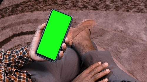 Black African male man sitting in swivel chair with crossed legs tapping leg with hand watching phone with green screen in room.