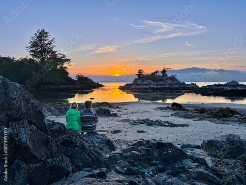 Couple sitting on beach watching sunset over the sea islands. Pacific Rim National Park. Broken Group Islands.  Tofino. British Columbia. Canada. 