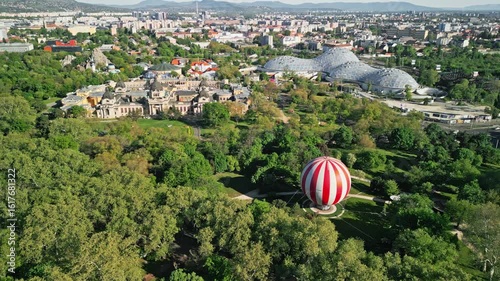 Fixed drone video from City Park center facing Széchenyi Thermal Bath, capturing the BalloonFly, Biodóm, and lush park surroundings in one wide morning frame in Budapest.  
