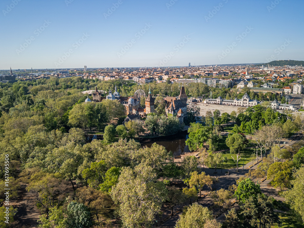 Fototapeta premium Wider aerial photo of Vajdahunyad Castle and artificial lake in Budapest, centered in lush City Park surroundings, with expanded view of greenery and park paths in soft morning light. 