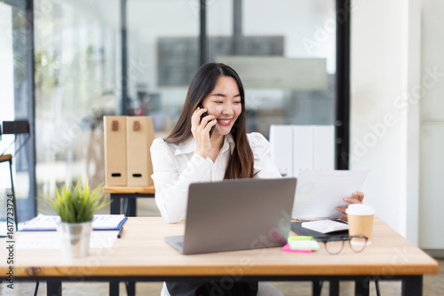 Asian woman talking on smartphone with laptop and project business planning in modern office.