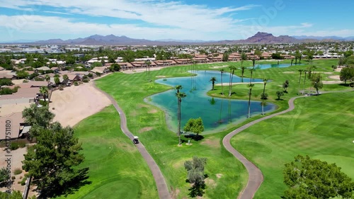 Aerial view of a green golf course with palm trees, lake, and homes under a clear blue sky