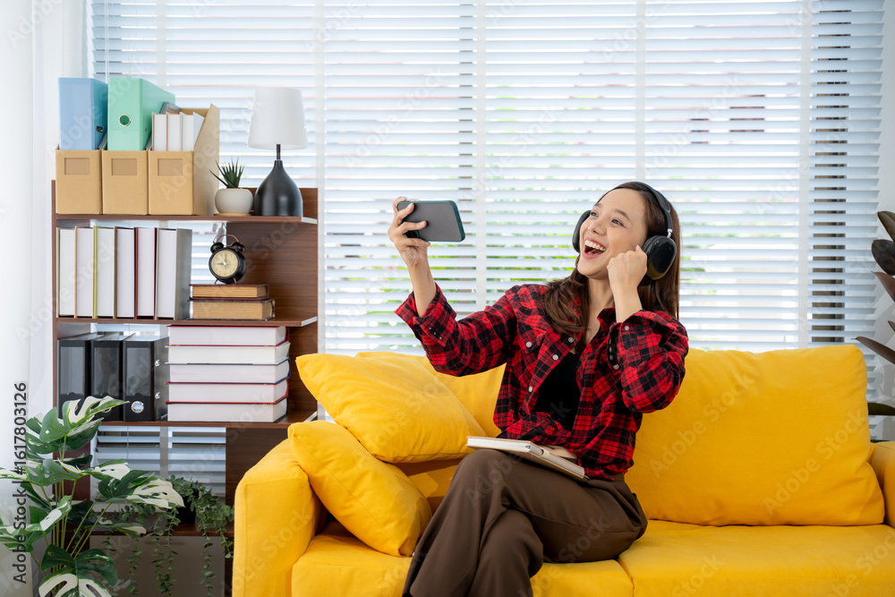 Obraz premium Cheerful Woman Enjoying Streaming Video: A joyful young woman wearing headphones sits on a bright yellow sofa, engrossed in a video streamed on her smartphone.