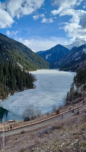 Frozen pale, icy blue-green mountain lake surrounded by majestic, snow-dusted peaks and dense pine forests under a bright, cloudy blue sky. Related to winter landscapes, mountain scenery, outdoor