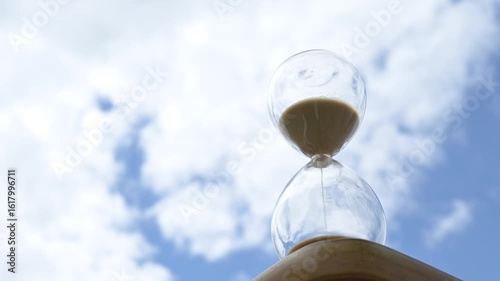 Glass hourglass against blue sky with clouds. Transparent hourglass filled with sand placed against a cloudy blue sky background. Concept of time, transience, waiting, and the passing of life