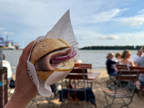 Traditional north german street food snack Fischbroetchen wheat bun with matjes fish fillet and onion in female hand close on Elbstrand beach