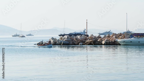 Wallpaper Mural Peaceful sunny morning in Varkiza, Greece. A small boat with two people leaves a wake on calm sea, yachts moored behind breakwater, distant mountains on the horizon Torontodigital.ca