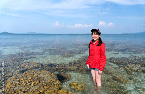 Young Asian girl wearing a red UV-protective long-sleeved shirt and cap stands in the shallow ocean surrounded by coral reefs on a sunny day, smiling at the camera. Koh Kradan, Trang, Thailand.