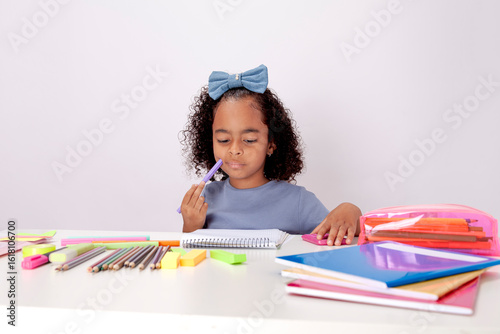 Fotografie Thoughtful schoolgirl, ethnic sitting at a table with school supplies attentive