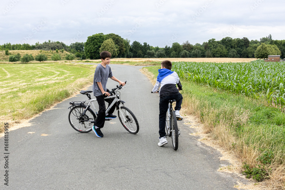 Obraz premium Two boys riding bicycles on a rural road surrounded by green fields and trees.